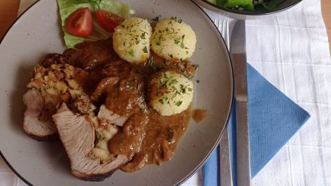 Ein Teller mit gefülltem Kalbsbraten, Knödeln und Soße, dekoriert mit Petersilie. Daneben eine Schüssel Salat.