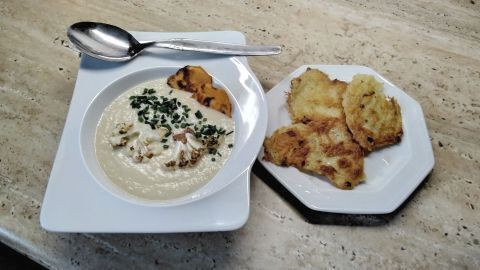 Eine Schale Blumenkohlcremesuppe mit Blumenkohlstücken, Süßkartoffelscheiben und Schnittlauch, daneben Kartoffelpuffer auf einem Teller.